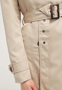 Beżowy płaszcz typu trench coat z szerokim kołnierzem, paskiem w talii, zatrzaskami i podwiniętymi mankietami, wykonany z gładkiej, teksturowanej tkaniny.