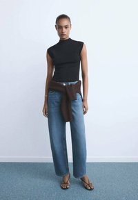 Zwarte mouwloze top gecombineerd met lichtblauwe wijde jeans, een donkerbruin truitje geknoopt om de taille, en zwarte sandalen met bandjes.