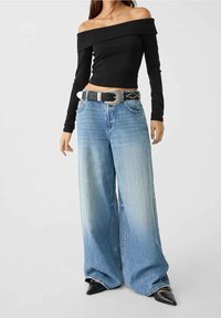 Top nero off-the-shoulder a maniche lunghe abbinato a jeans blu sbiaditi a gamba larga, accentuato da una cintura nera con fibbia argentata e decorazioni.
