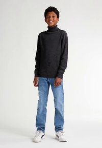 Pulover negru cu guler înalt din material tricotat, asortat cu blugi albastru deschis și adidași albi. Modelul stă pe un fundal neutru.