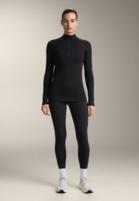 Fekete, hosszú ujjú sportfelső, negyedcipzárral, sima textúrával; fekete leggingszel párosítva. A modell fehér zoknit és futócipőt visel.