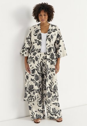 Kobieta z kręconymi włosami, ubrana w luźne, kremowe kimono w kwiaty i pasujące szerokie spodnie na białą bluzkę i sandały.