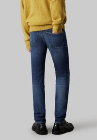 Džínsy v tmavo modrej farbe so slim fit strihom, s uhlovými zadnými vreckami, nosené s štruktúrovaným žltým sveterom a čiernymi topánkami.