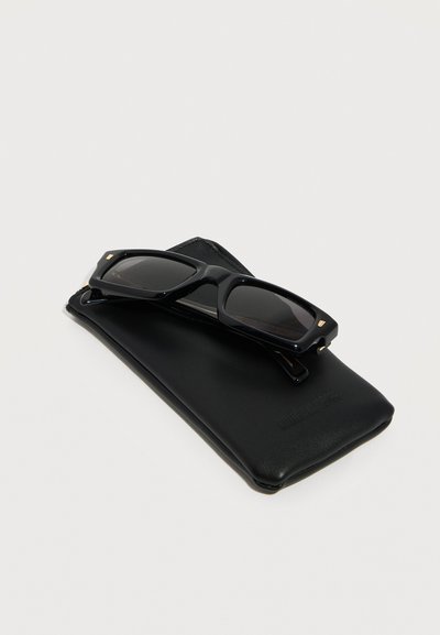 Μαύρα ορθογώνια γυαλιά ηλίου που ακουμπούν σε μαύρη δερμάτινη θήκη πάνω σε απλό ανοιχτόχρωμο φόντο.