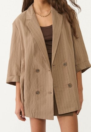 Femme portant un blazer beige à fines rayures oversize avec les manches retroussées, sur un top et une jupe marron, accessoirisé d'un collier chaîne doré.
