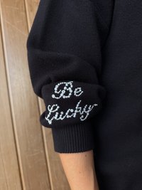 Czarny dzianinowy sweter z prążkowanymi mankietami oraz napisem „Be Lucky” zdobionym srebrnymi cekinami na lewym rękawie. Gładka tekstura i dopasowany fason.