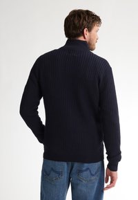 Marine gebreide trui met hoge kraag, geribbelde textuur en lange mouwen, gecombineerd met blauwe jeans met stiksels op de achterzakken.