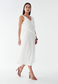 Weißes, ärmelloses Kleid mit Rüschendetails, Textur und tiefem V-Ausschnitt. Paar tanfarbene Ankle-Strap-Absätze und lange, glatte Haare. Schlichter Hintergrund.
