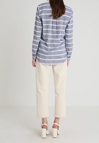 Sininen raidallinen pitkähihais Shirt takana olevan laskoksen kanssa, yhdistettynä kermaisiin leveälahkeisiin housuihin, joissa on rispaantuneet lahkeet. Yhdistettyinä valkoisten paksupohjaisten sandaalien kanssa.