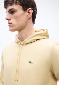 Jeune homme en sweat à capuche beige avec un logo de crocodile vert, regardant vers la gauche sur un fond blanc uni.