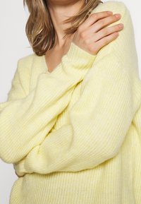 Weicher, übergroßer gelber Pullover mit V-Ausschnitt, gerippter Textur und langen Ärmeln. Der Stoff wirkt leicht und gemütlich, mit subtilen Farbvariationen.