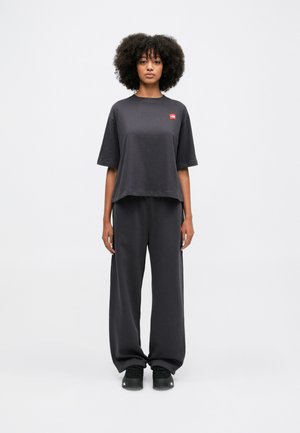 Tumši pelēki kokvilnas brīvā stila T-krekls ar mazu sarkanu logo, savienots ar atbilstošām brīvā stila biksēm un melniem sporta apaviem.