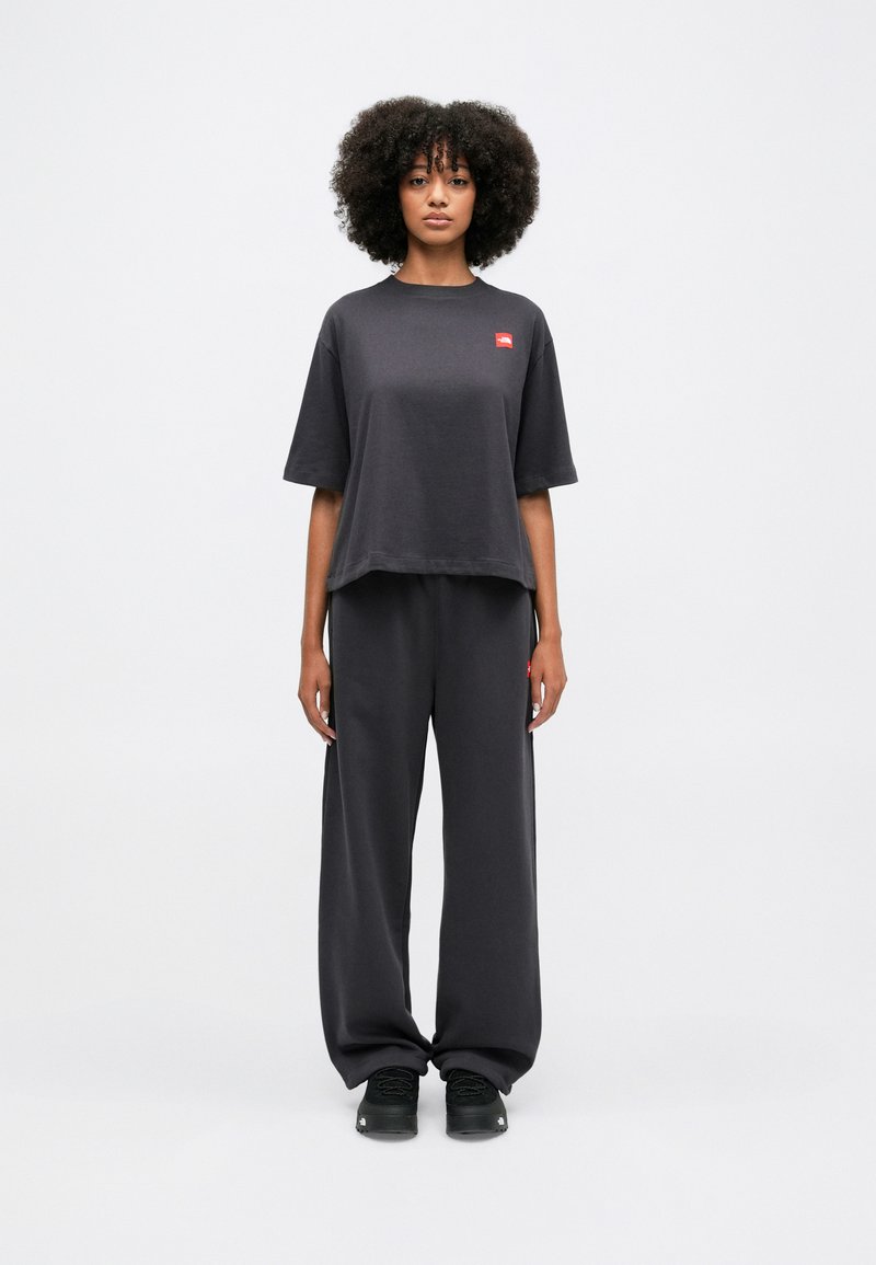 T-shirt oversize grigio scuro in cotone con un piccolo logo rosso, abbinato a pantaloni larghi coordinati e sneakers nere.