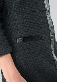 Prenda texturizada negra con diseño de cuello, que presenta acentos negros brillantes y cierre de botones, resaltada por un elegante detalle en el bolsillo.