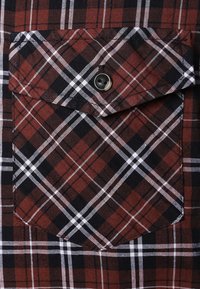 Gros plan d'une poche de chemise à carreaux rouges, noirs et blancs avec un rabat fixé par un seul bouton noir.