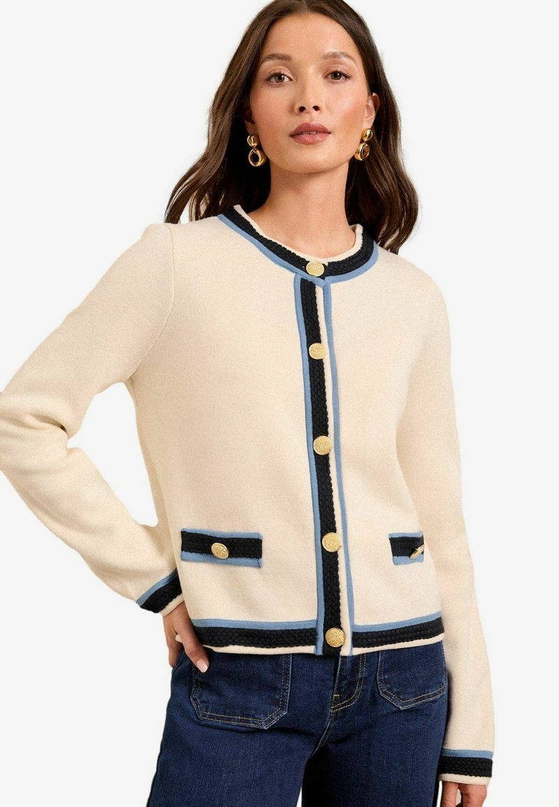 Beige neulecardigan, jossa on musta ja sininen reunus, kultaiset napit ja kaksi etutaskua, valmistettu pehmeästä, teksturoidusta kankaasta.