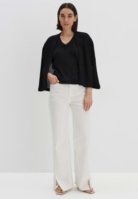 Schwarzes geripptes Stricktop mit V-Ausschnitt und offenen Ärmeln, kombiniert mit weißen ausgestellten Hosen mit seitlichen Schlitzen, getragen mit beigen flachen Sandalen.