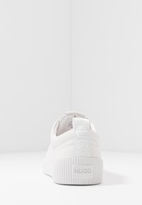 Baskets blanches avec une tige texturée, une semelle plate et un passant de talon marqué. Présente un design minimaliste et une finition lisse.