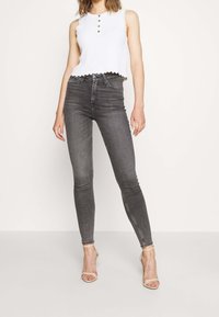 Top blanco sin mangas con bordes festoneados y tres botones, combinado con jeans ajustados de cintura alta en gris oscuro y tacones desnudos con tiras.