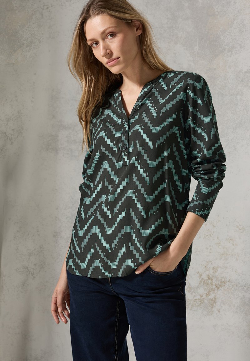 Blusa nera con motivo a zigzag turchese, maniche lunghe e scollo a V. Tessuto liscio con una leggera lucentezza, dotata di una patta con tre bottoni.