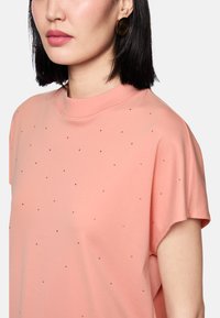 Short-sleeve, peach-colored top with a crew neck, featuring small gold studs evenly distributed across the fabric for added detail.