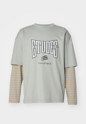 Vaaleanharmaa ylisuurikokoinen t-paita lyhyillä hihoilla, jossa on rohkea "ETUDES PARIS" -logo, kerroksittain pitkähihaisen beige- ja raidallisen paidan päällä.