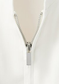 Makro záber na strieborný kovový zips čiastočne zapnutý na bielom textúrovanom látke, zobrazujúci detail zubov zipsu a ťahadla.