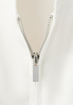 Prim-plan cu un fermoar metalic argintiu parțial închis pe o țesătură albă texturată, evidențiind detaliile dinților fermoarului și a trăgătorului.