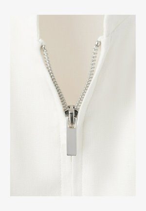 Gros plan sur une fermeture éclair en métal argenté partiellement fermée sur un tissu blanc texturé, montrant en détail les dents de la fermeture éclair et la languette de tirage.