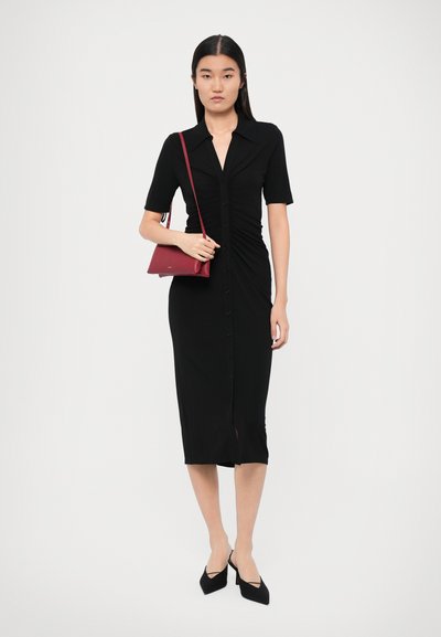 Robe midi noire avec des manches courtes, devant boutonné et détails froncés. Le modèle tient un sac à main rouge, portant des escarpins noirs à bout pointu.