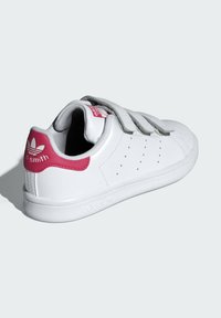 Baskets blanches en cuir avec une sangle Velcro grise, un œillet de talon rose, des détails perforés et une semelle en caoutchouc texturée.