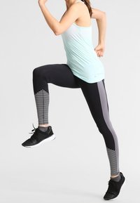 Leggings de sport noirs et gris avec un motif à rayures sur le bas de la jambe, portés avec un débardeur bleu clair et des chaussures de sport noires.