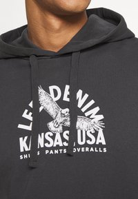 Felpa grigia con cappuccio e cordino, con una grafica bianca di un'aquila e la scritta "LEVI'S DENIM KANSAS, USA" in lettere grasse. Materiale in cotone.
