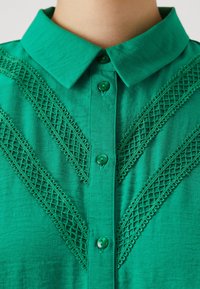 Blusa verde con colletto, caratterizzata da dettagli in pizzo testurizzato a motivo incrociato e bottoni verdi lungo la parte anteriore. Tessuto morbido.