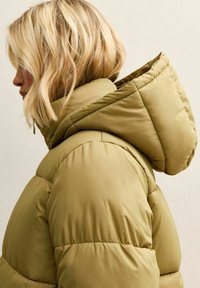 Chaqueta acolchada de color verde oliva con capucha, presenta una textura acolchada y un ajuste holgado. El material parece suave y ligero.