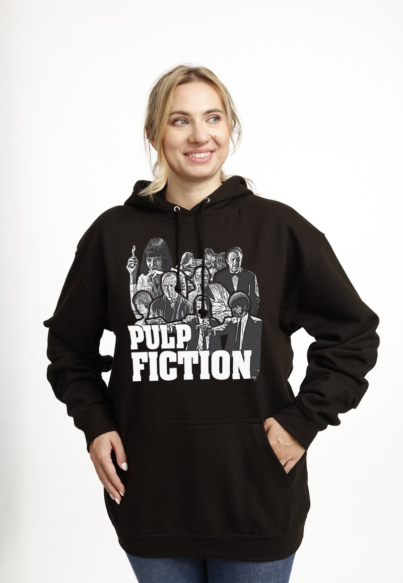 Donna sorridente con felpa nera con cappuccio con disegno frontale in bianco e nero dei personaggi e del titolo del film Pulp Fiction, mani nelle tasche.