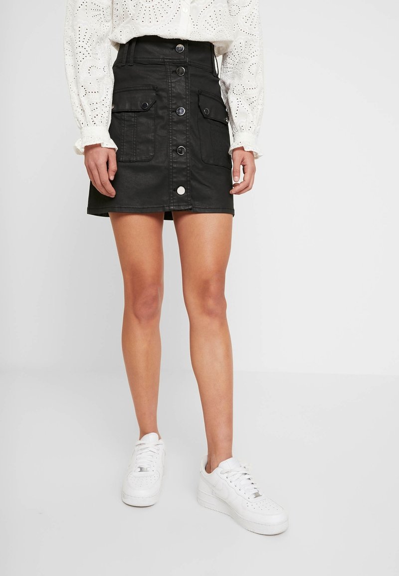 Svart denim mini kjol med framknäppning, två sidofickor och en figurnära silhuett. Bärs med en vit mönstrad blus och vita sneakers.