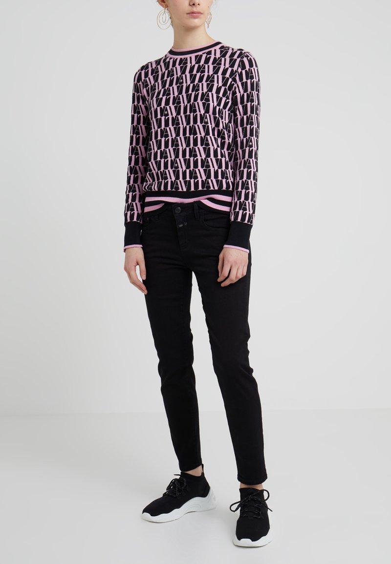 Personne portant un pull à motifs rose et noir avec un pantalon noir et des baskets noires à semelles blanches, debout devant un fond uni.