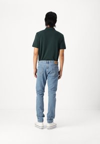 Muž stojící zády, oblečený v tmavě zeleném polo tričku, světlých modrých džínech a bílých teniskách Converse na bílém pozadí.