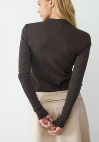 Langärmliges braunes Oberteil aus glattem Stoff, mit einer figurbetonten Passform und weicher Textur. Getragen mit einem beigen Rock, die Hände hinten verschränkt.