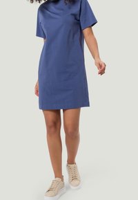 Femme marchant, portant une robe bleue courte et des baskets à plateforme beiges sur un fond léger uni.