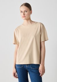 Beige Baumwoll-T-Shirt mit kurzen Ärmeln und Rundhalsausschnitt, mit einem lässigen Schnitt. Getragen mit blauen Jeans.