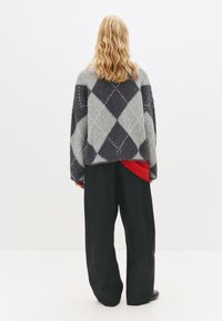 Pulover din lână supradimensionat, cu un model argyle gri și negru, cu un finisaj texturat, purtat peste un top roșu, asortat cu pantaloni largi negri.