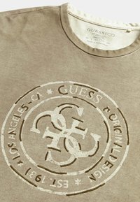Šviesiai rudas medvilninis marškinėlis su įspaustu apvaliu logotipu, kuriame yra tekstas "GUESS" ir "EST. 1981" kontrastinga kreminė spalva.
