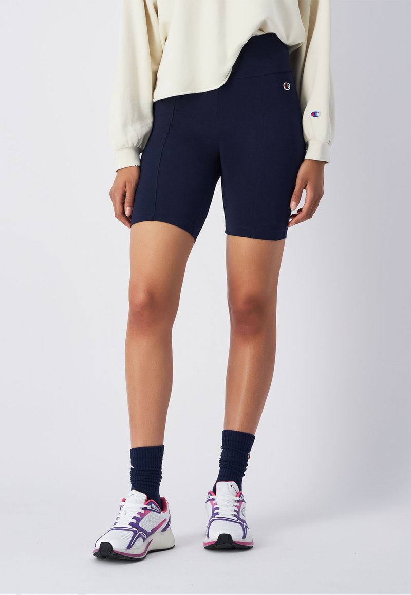 Shorts de cyclisme taille haute bleu marine en tissu extensible, avec une texture lisse et un logo sur le côté. Associés à des chaussures de sport blanches et des chaussettes bleu marine.