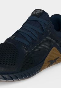 Κοντινό πλάνο ενός αθλητικού παπουτσιού Reebok σε σκούρο μπλε με διχτυωτή υφή, μπλε κορδόνια και ανοιχτό καφέ σόλα με το λογότυπο της Reebok.