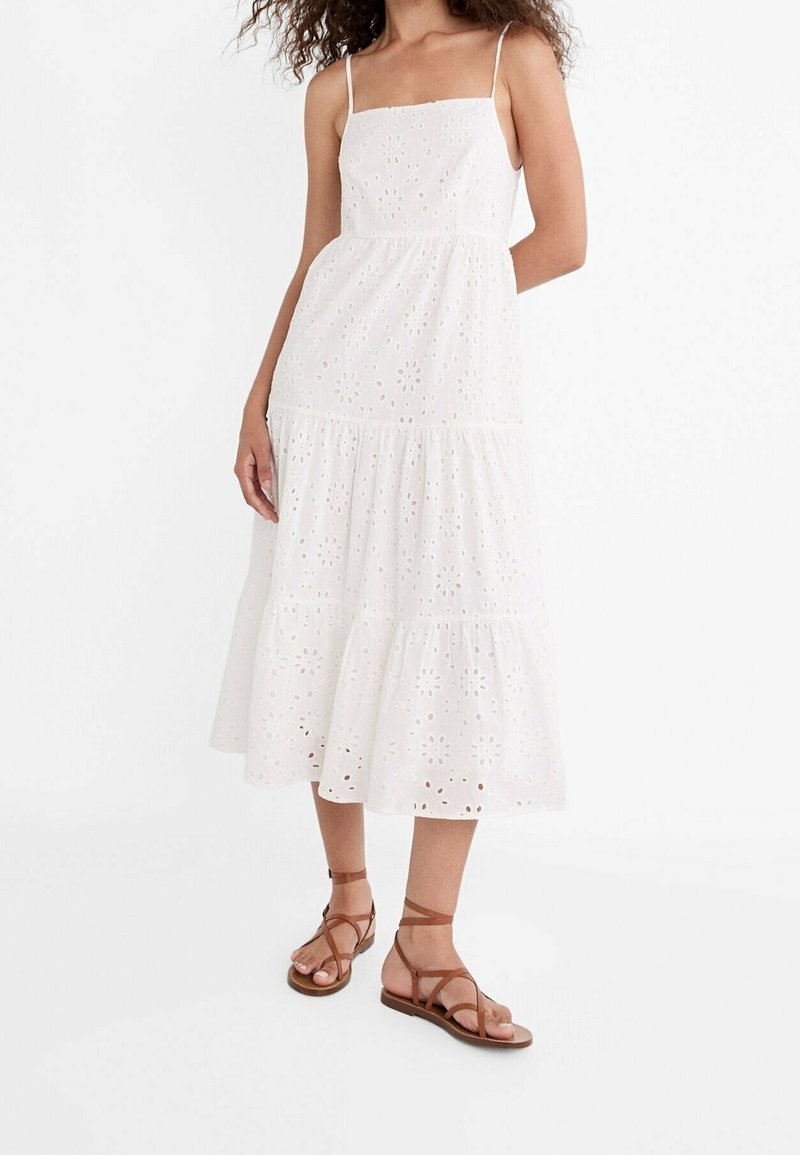 Femme portant une robe midi blanche sans manches à œillets avec de fines bretelles et des sandales plates marron à brides sur fond blanc.