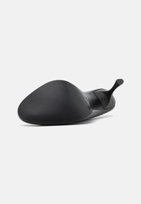 Svart stilettohögklackad sko sedd från sulan, med spetsig tå och tunn kurvad klack mot en vit bakgrund.