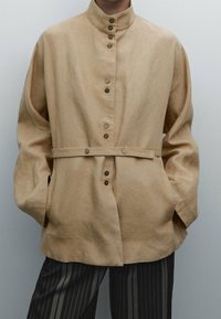 Beige nappatakki korkealla kauluksella, pitkät hihat ja etutaskut; sisältää vyötärösuunnittelun, jossa on metallinappeja. Pintatekstuuri näyttää pehmeältä.