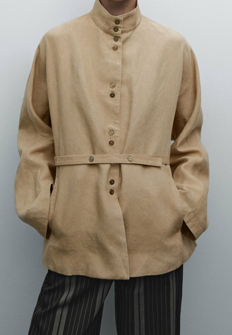 Beige nappatakki korkealla kauluksella, pitkät hihat ja etutaskut; sisältää vyötärösuunnittelun, jossa on metallinappeja. Pintatekstuuri näyttää pehmeältä.